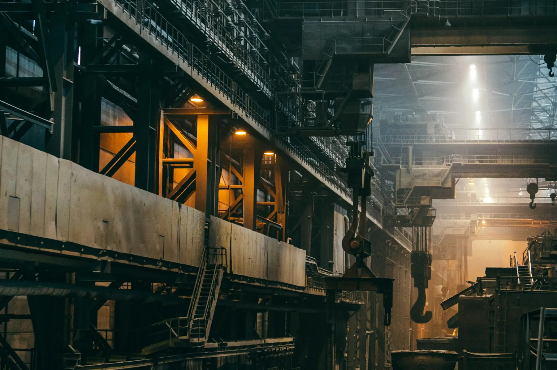 Interior of a large heavy-industry plant with overhead steel structures, crane hooks and machinery in low light